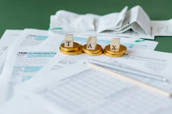 Pile of tax forms with gold coins spelling tax and a pen on green surface