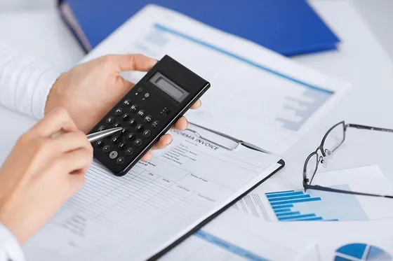 Hands using a calculator and pen to review financial documents with charts and a blue binder on a desk.
